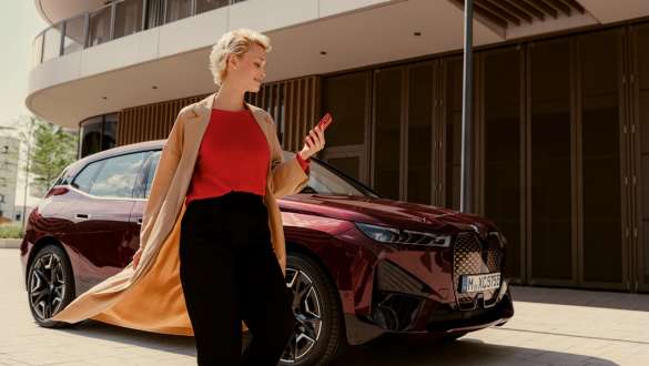 My BMW App My BMW App ผู้หญิงยืนอยู่บนถนนถือสมาร์ทโฟนไว้ในมือและมีรถยนต์ BMW อยู่ด้านหลัง